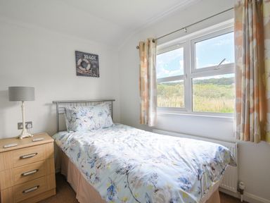 A bedroom with a single bed next to a wooden nightstand with a lamp and a window with floral curtains at The Hideaway in Eype