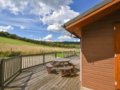 A wooden deck with a round picnic table and benches next to a brown wooden building surrounded by grass and hills at The Hideaway in Eype