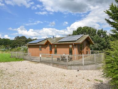 A single story wooden house with solar panels on the roof and a fenced wooden deck surrounded by trees and grass at The Hideaway in Eype