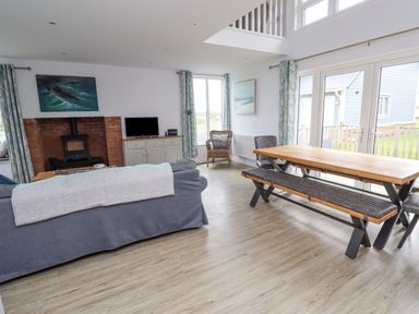 A living room with a gray sofa a wooden dining table with bench and chairs a fireplace and large windows at Turning Tide in Beadnell