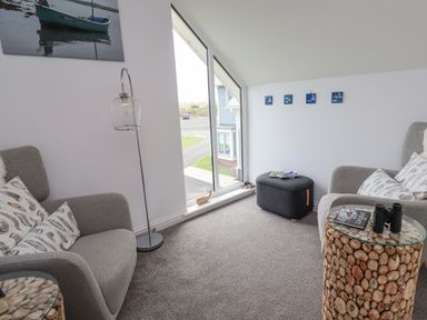 A sitting area with two grey armchairs a floor lamp a glass topped side table and a small stool by a window at Turning Tide in Beadnell