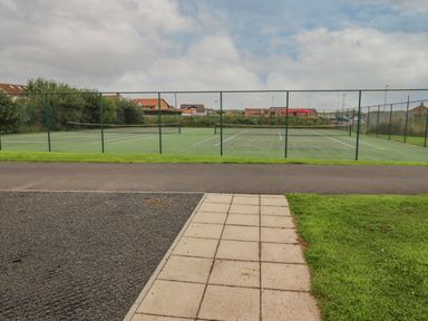 Two tennis courts surrounded by a green chain link fence with a paved path and grass in the foreground at Turning Tide in Beadnell