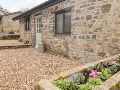 A stone building with a white door and a window and a flower bed with pink and purple flowers in front at The Stable in Amroth