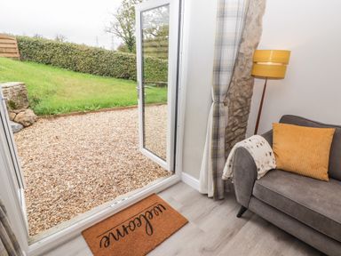 An open glass door leading to a gravel outdoor area with grass and a hedge inside a room with a grey sofa yellow cushion and floor lamp at The Stable in Amroth