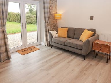 A sitting room with a grey sofa and yellow cushions next to a small wooden side table and glass doors with curtains at The Stable in Amroth