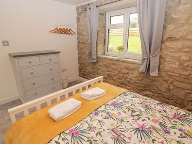 A bedroom with a bed covered by a floral bedspread and yellow blanket next to a window with curtains and a grey chest of drawers at The Stable in Amroth