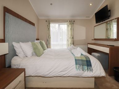 A bedroom with a bed with green pillows and folded towels a window with curtains a wall-mounted television and a dresser with a mirror at Haven Lodge in Bossiney Bay near Tintagel