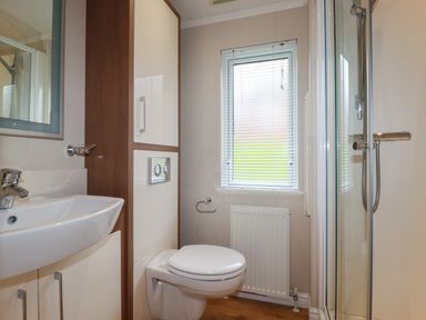 A bathroom with a sink, toilet, shower, window with blinds, and storage cabinet at Haven Lodge Bossiney Bay near Tintagel