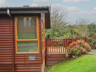 A wooden lodge with a glass window and a wooden deck with railing in a green garden at Haven Lodge Bossiney Bay near Tintagel