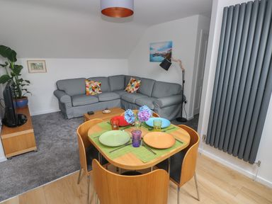 A living room with a gray corner sofa and a wooden dining table with four plates and flowers at The Loft @ Wisteria Cottage in Saundersfoot