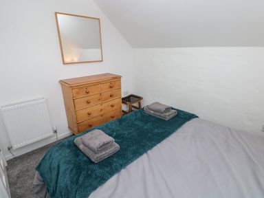 A bedroom with a bed covered in teal and gray bedding a wooden chest of drawers with a mirror above and a small stool at The Loft @ Wisteria Cottage in Saundersfoot