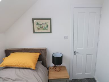 A bedroom corner with a bed a yellow pillow a bedside table with a lamp a framed picture on the wall and a closed white door at The Loft @ Wisteria Cottage in Saundersfoot