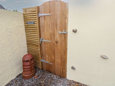 A wooden gate with a latch and sign The Loft next to a cream wall with embedded stones and a red vent on gravel at The Loft @ Wisteria Cottage in Saundersfoot