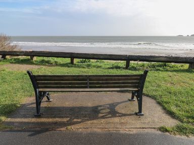 A wooden bench on a paved area facing the sea with a grassy area and wooden railing behind at The Loft @ Wisteria Cottage in Saundersfoot