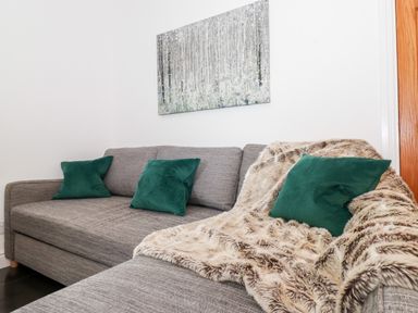A grey fabric sofa with green cushions and a beige fur throw in a room with white walls and a forest painting at Watersong in Coniston