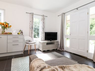 A living room with a television on a stand a white door curtains sofa with a fur throw and a kitchen counter with flowers at Watersong in Coniston