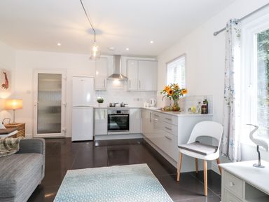 A kitchen with white cabinets a white refrigerator a gray sofa a chair with cushion and a window with floral curtains at Watersong in Coniston
