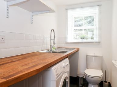 A laundry room with a washing machine under a wooden countertop a stainless steel sink a toilet and a window with blinds at Watersong in Coniston