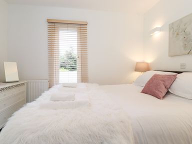 A bedroom with a bed with white linens pink cushion and folded towels near a window with blinds and a dresser with a mirror at Watersong in Coniston
