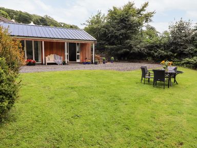 A lawn with outdoor table and chairs with flowers near a wooden house with a gravel area and trees at Watersong in Coniston