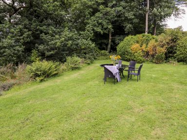 A lawn with a round glass table and black chairs surrounded by trees and bushes at Watersong in Coniston
