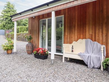 An outdoor patio area with a wooden bench covered with a blanket and a bottle with glasses near a wooden wall and potted plants