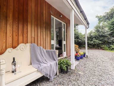 An outdoor porch with a cream bench covered with a gray blanket a bottle of wine and a wine glass next to potted plants and flowers at Watersong in Coniston