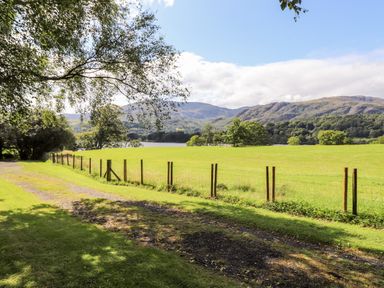 A grassy field with a wire fence and a dirt path beside trees and hills at Watersong in Coniston