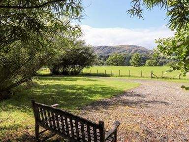 A wooden bench on grass near trees overlooking a field with mountains in the background at Watersong in Coniston