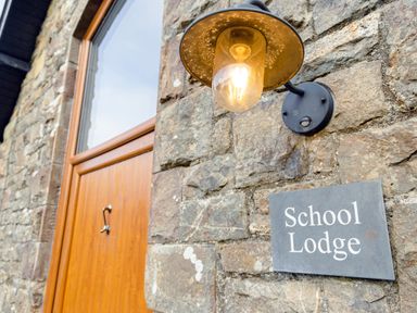 A stone wall with a wooden door a light fixture and a sign that reads School Lodge in Llandeilo
