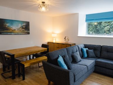 A living room with a wooden dining table and black chairs a gray sectional sofa with blue and gray cushions and a wooden sideboard with a lamp and decoration at School Lodge in Llandeilo