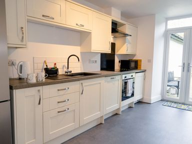 A kitchen with white cabinets a black sink a kettle two mugs an oven a microwave and glass doors at School Lodge in Llandeilo