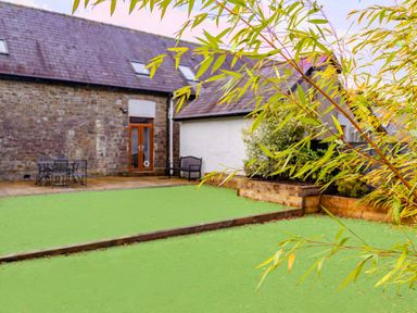 A backyard with green artificial turf, a stone wall house, patio with outdoor furniture, and plants in School Lodge in Llandeilo