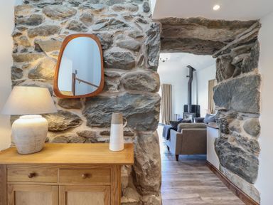 A wooden cabinet with a lamp and vase beside a stone wall opening leading to a living room with a stove at Gaerau Bach in Llanaber