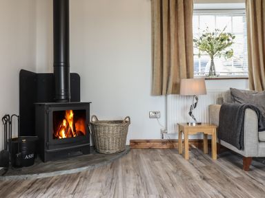 A wood burning stove with fire a basket and fire tools next to a window with curtains a lamp on a wooden side table and part of a sofa at Gaerau Bach in Llanaber
