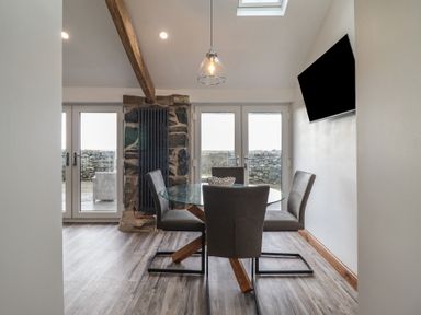 A dining area with a round glass table and four chairs under a hanging light with a wall-mounted TV and glass doors at Gaerau Bach in Llanaber