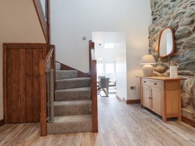 A hallway with carpeted stairs and a wooden sideboard with a lamp and mirror on a stone wall at Gaerau Bach in Llanaber