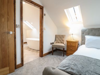 A bedroom with a bed chair nightstand and lamp next to a doorway showing a bathroom with a bathtub at Gaerau Bach in Llanaber