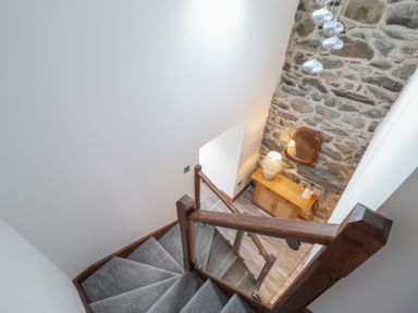 A staircase with grey carpet and wooden railing overlooking a small cabinet with a lamp and mirror against a stone wall at Gaerau Bach in Llanaber