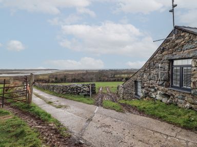 A stone building beside a concrete and dirt driveway with a gate and fields in the background at Gaerau Bach in Llanaber