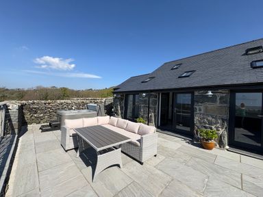 An outdoor patio with a stone floor a rattan sofa set and a hot tub next to a stone building at Gaerau Bach in Llanaber