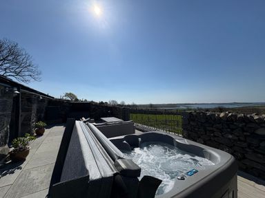 A patio with a hot tub seating area potted plants and stone walls overlooking a field and water at Gaerau Bach in Llanaber