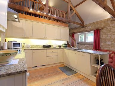 A kitchen with cream cabinets wooden beams a granite countertop and a window with red curtains at The Granary in Bourton-On-The-Water