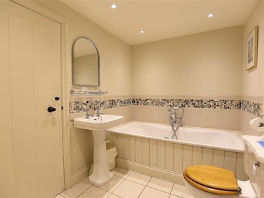 A bathroom with a pedestal sink and mirror a bathtub with mosaic tile border and a toilet with wooden seat at The Granary in Bourton-On-The-Water