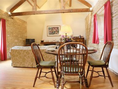 A dining area with wooden chairs and table set with plates and glasses next to a living room with sofas and a television at The Granary in Bourton-On-The-Water