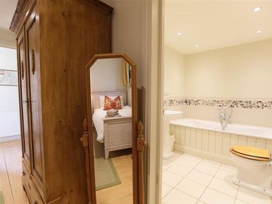 A bedroom with a wooden wardrobe and a mirror reflecting a single bed next to a bathroom with a bathtub and toilet at The Granary in Bourton-On-The-Water