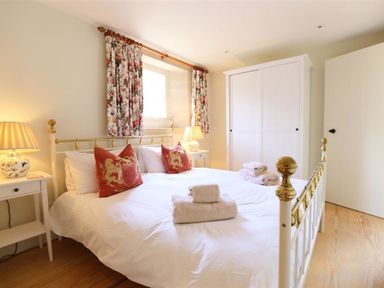 A bedroom with a double bed red pillows two nightstands with lamps a window with floral curtains and a white wardrobe at The Granary in Bourton-On-The-Water