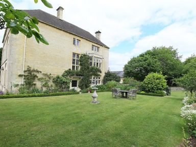 Exterior of a stone house with a large lawn garden table and chairs and trees at The Granary in Bourton-On-The-Water