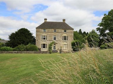 A three-story stone house with a pitched roof surrounded by trees and grass at The Granary in Bourton-On-The-Water