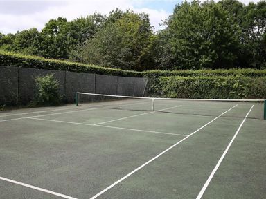 An outdoor tennis court with a net surrounded by green trees and hedge at The Granary in Bourton-On-The-Water